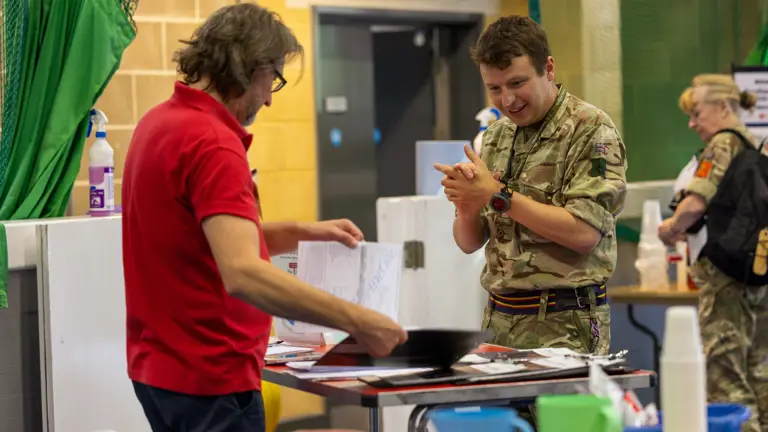 A man in a red shirt reviews documents while a soldier in camouflage listens attentively indoors.