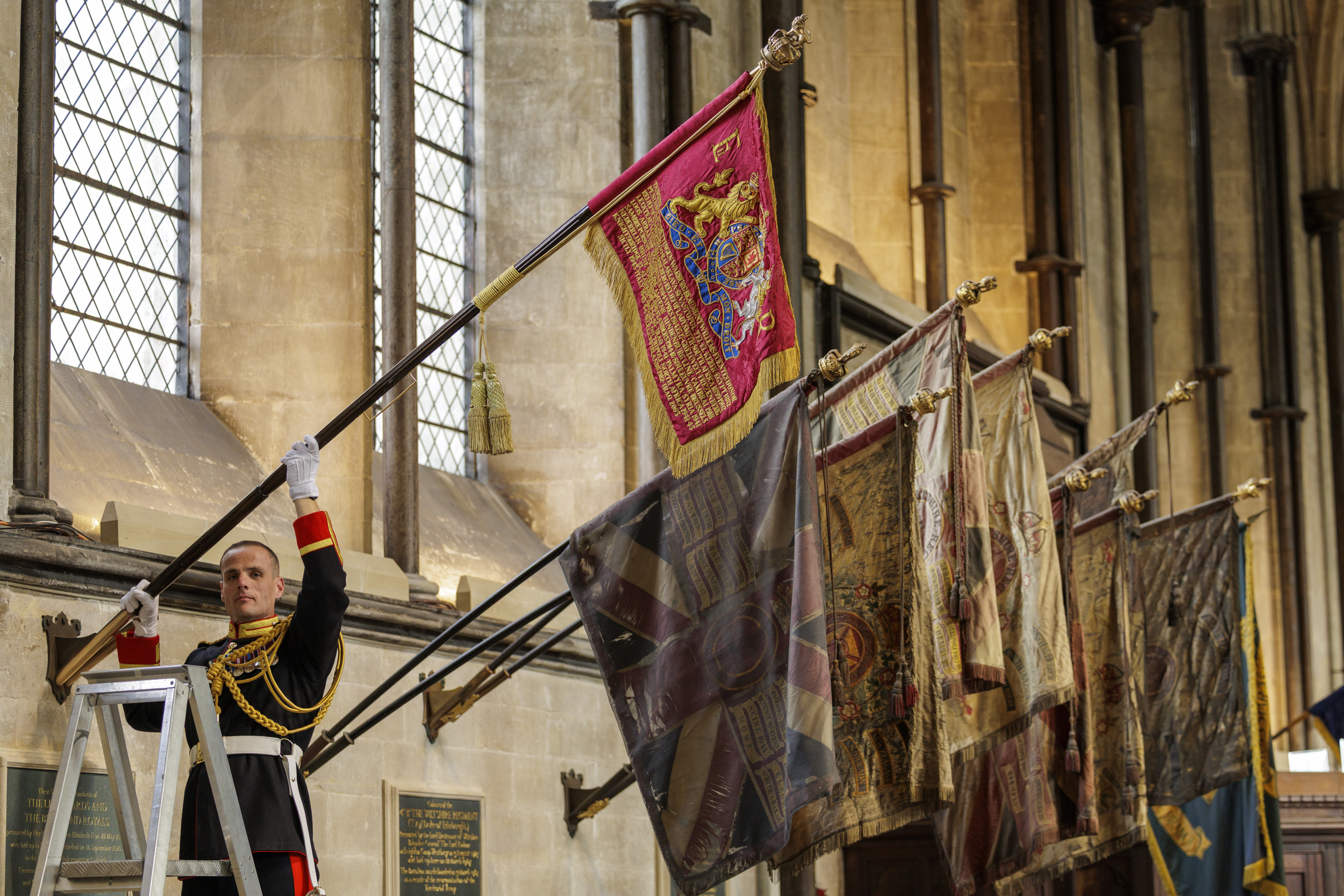 A uniformed soldier in ceremonial dress raises a red flag with gold embroidery inside a historic stone building with tall windows.