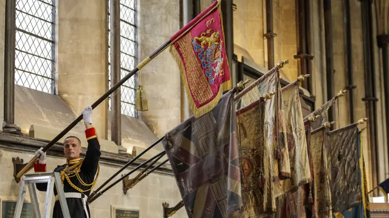 A uniformed soldier in ceremonial dress raises a red flag with gold embroidery inside a historic stone building with tall windows.