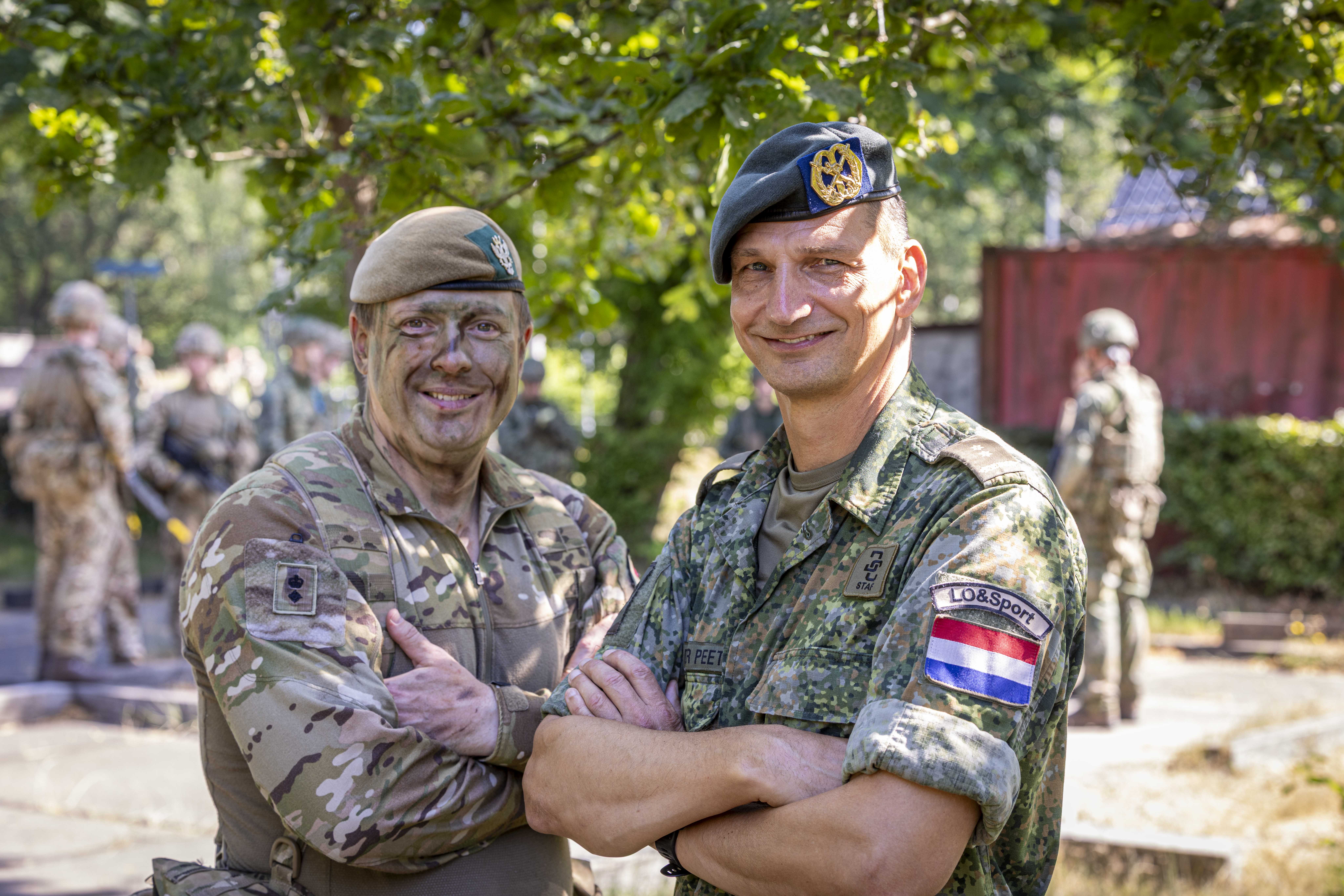 Two individuals in military uniforms stand smiling with arms crossed. One wears camouflage with a tan beret and has painted face, while the other wears a green beret with a badge, showing the Dutch flag patch and the word 