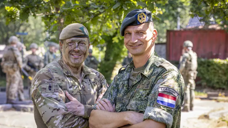 Two individuals in military uniforms stand smiling with arms crossed. One wears camouflage with a tan beret and has painted face, while the other wears a green beret with a badge, showing the Dutch flag patch and the word