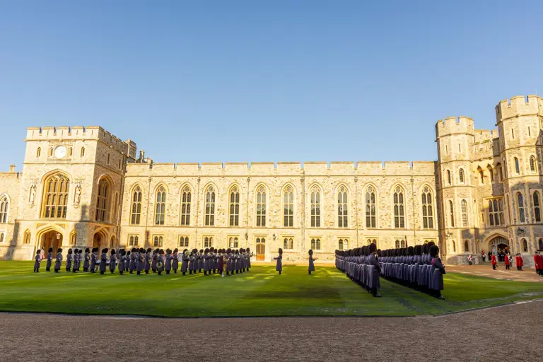 Ceremonial guards in uniforms stand in organised rows on a green lawn in front of a large, historic building under a clear blue sky, creating a formal atmosphere.
