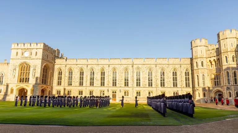 Ceremonial guards in uniforms stand in organised rows on a green lawn in front of a large, historic building under a clear blue sky, creating a formal atmosphere.