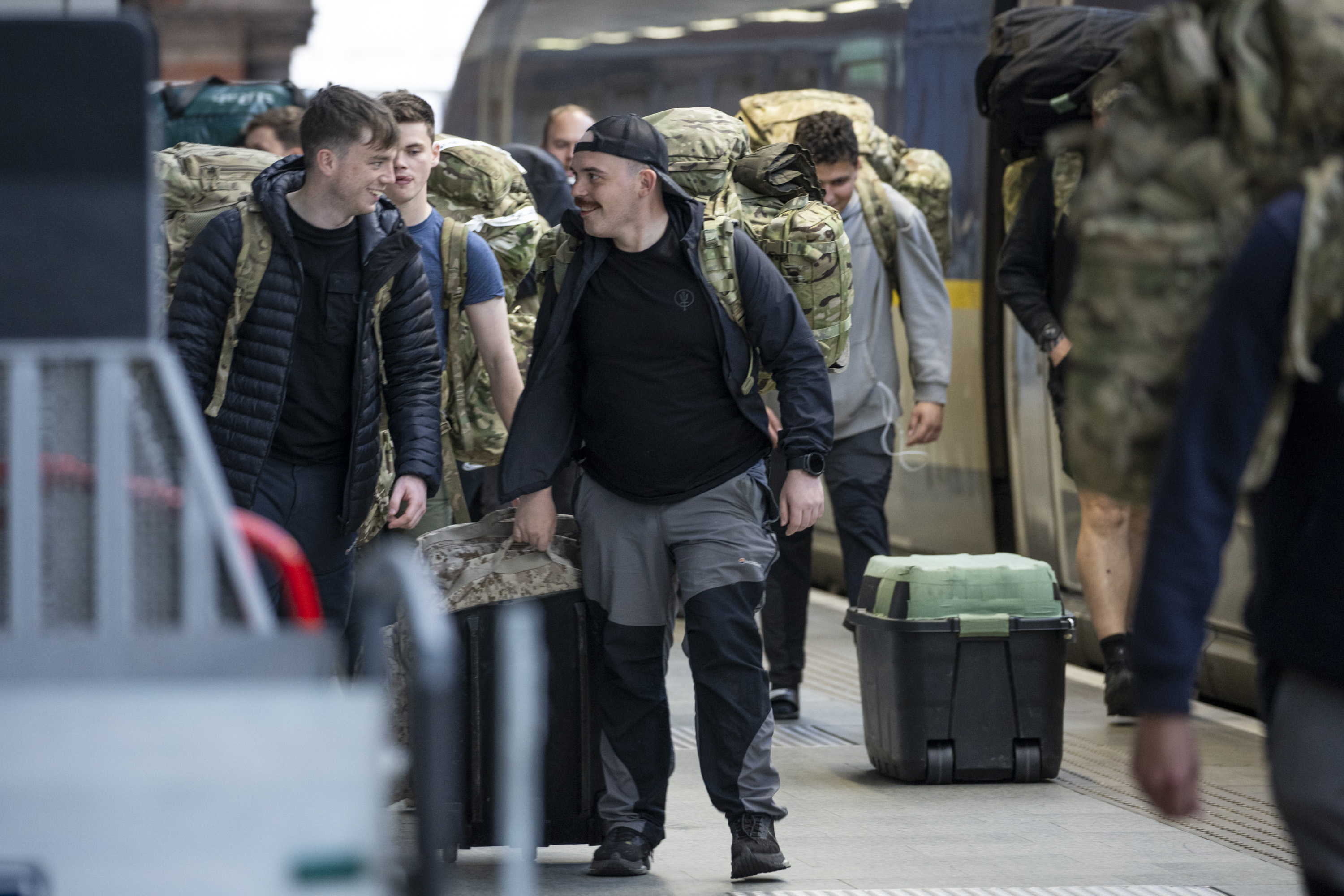 Men carry camouflage bags while walking along a train platform.