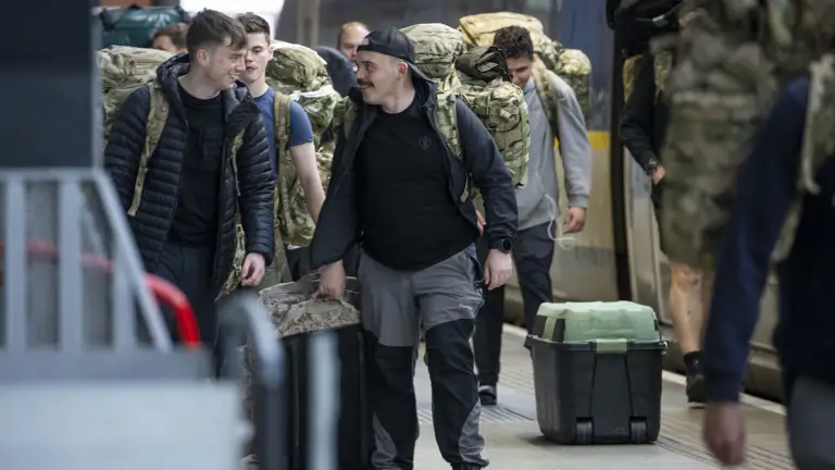 Men carry camouflage bags while walking along a train platform.