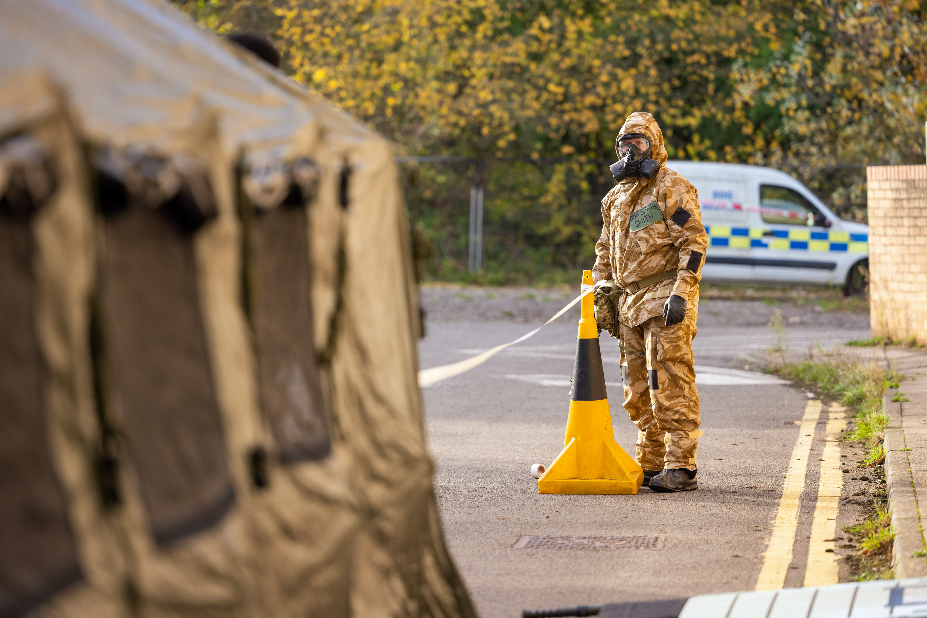 A soldier in a camouflage hazmat suit stands by a cordon.