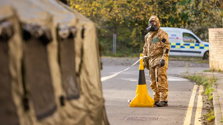 A soldier in a camouflage hazmat suit stands by a cordon.