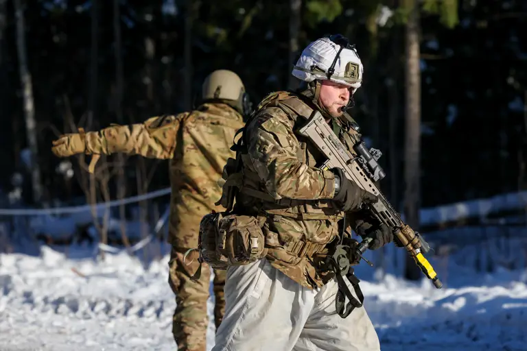 Soldier in winter camouflage holding a rifle with a yellow muzzle cover in a snowy forest setting.