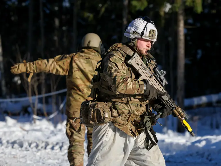 Soldier in winter camouflage holding a rifle with a yellow muzzle cover in a snowy forest setting.
