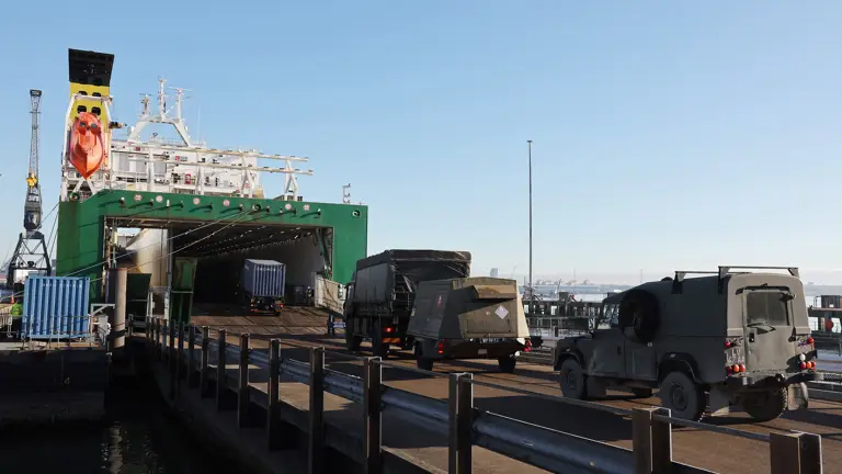 Military vehicles are shown being loaded onto a ship in convoy ready for transportation.