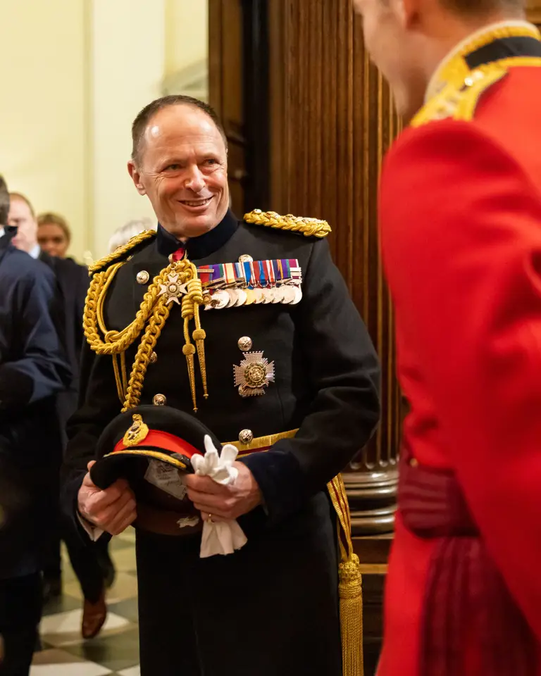 A military officer in a decorated black uniform holding a ceremonial hat and white gloves indoors.