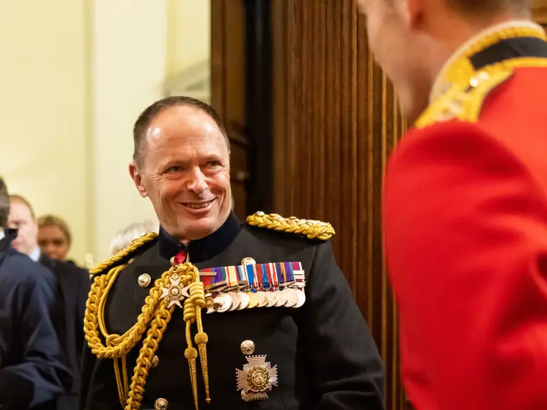 A military officer in a decorated black uniform holding a ceremonial hat and white gloves indoors.