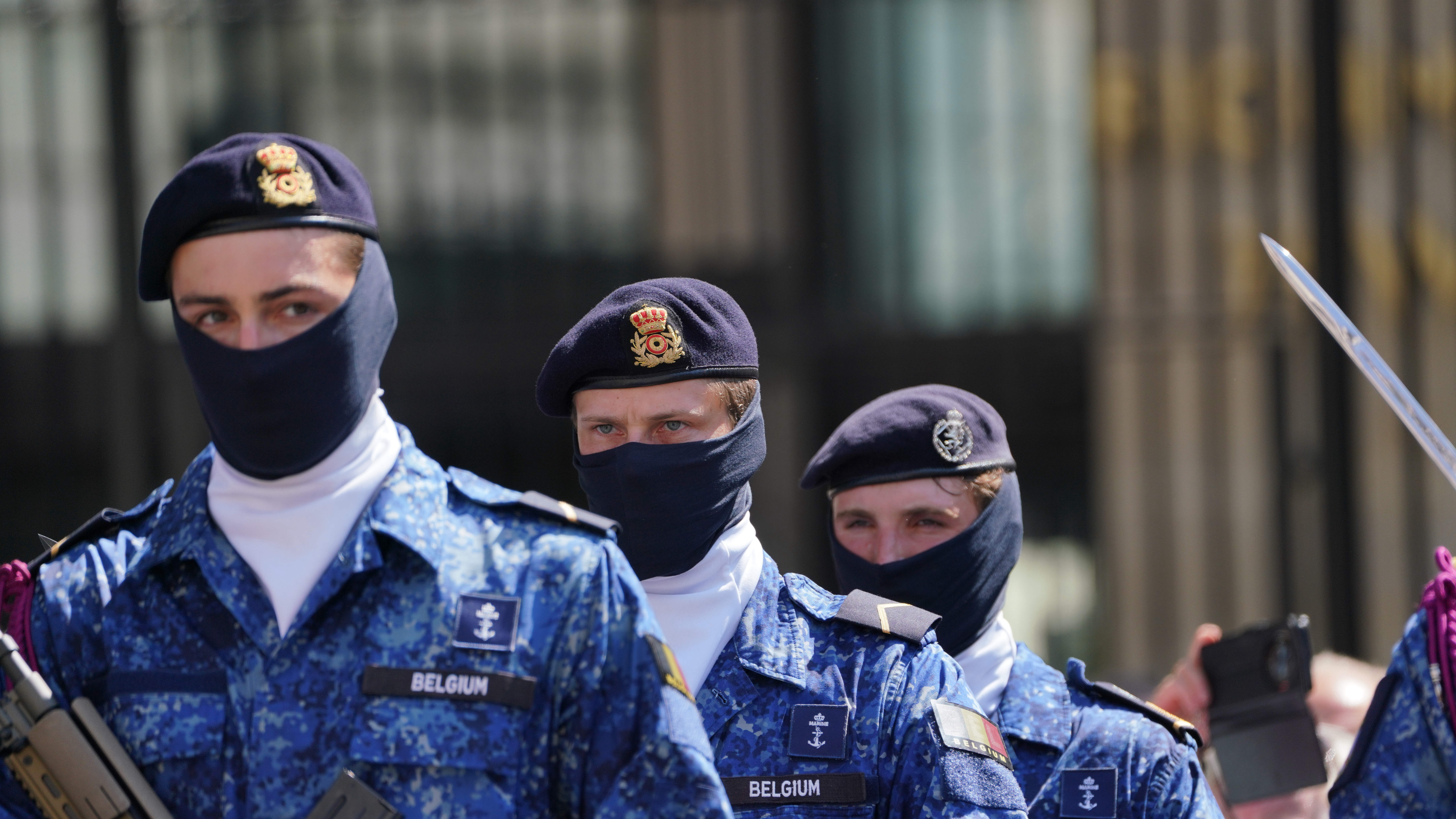 Belgian soldiers dressed in blue camouflage uniforms and berets, face coverings visible, stand together holding rifles. 