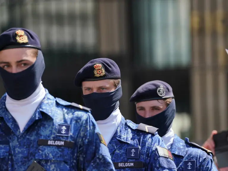 Belgian soldiers dressed in blue camouflage uniforms and berets, face coverings visible, stand together holding rifles.