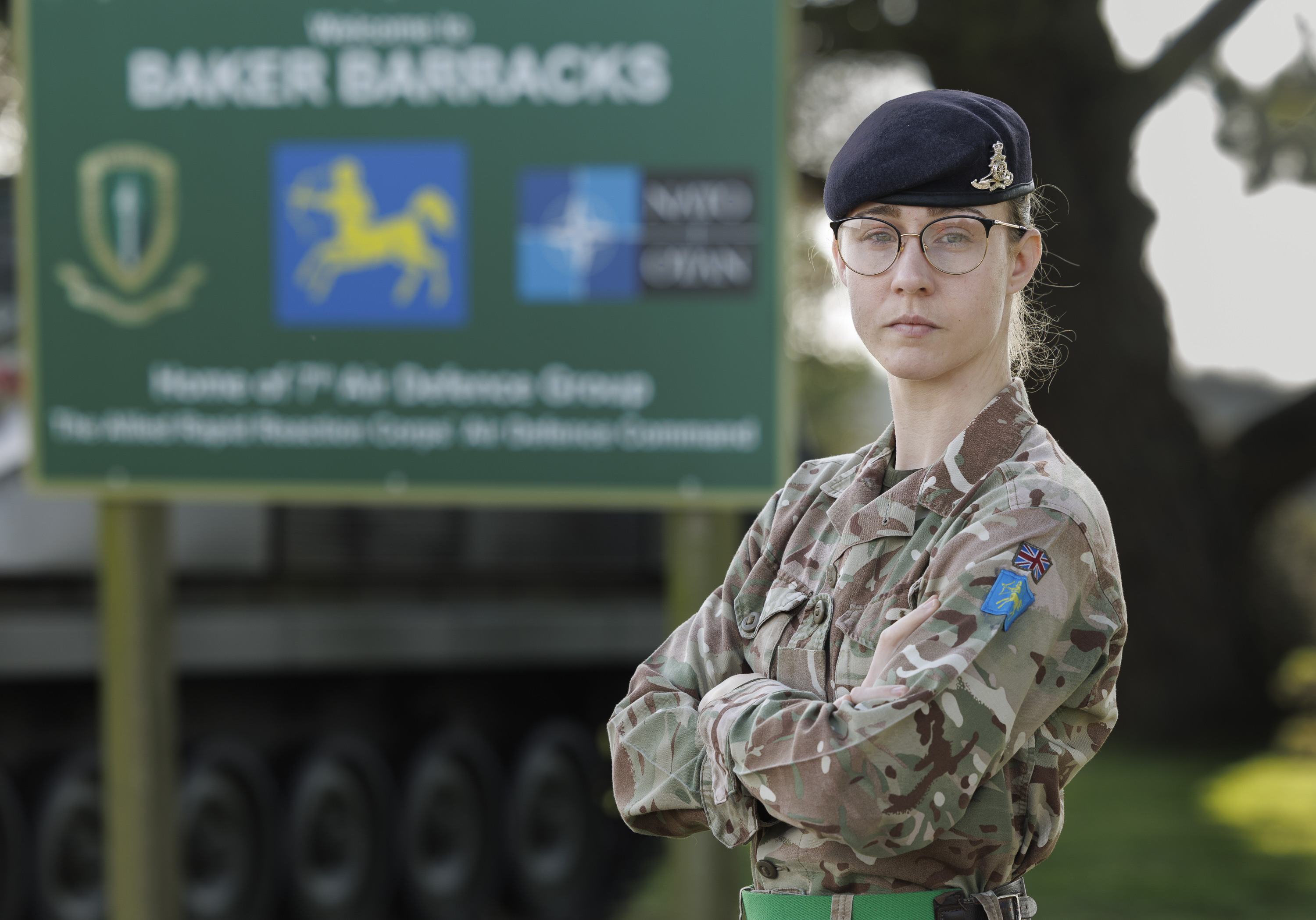 An individual in military camouflage stands confidently with arms crossed in front of a military vehicle equipped with missiles. The background includes grass and trees.