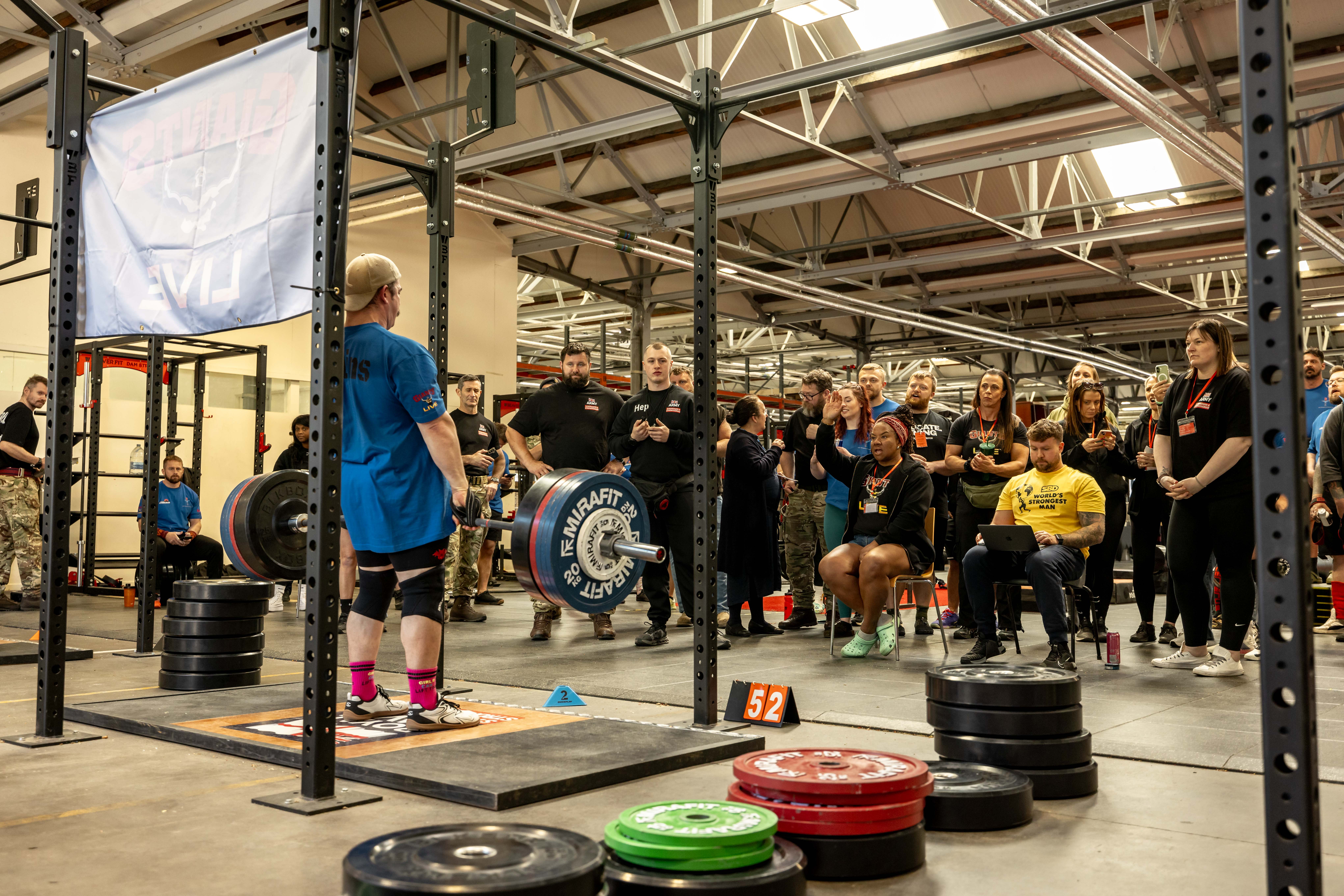 A powerlifter prepares to deadlift in a gym, surrounded by a focused crowd. Weight plates and equipment create an intense, competitive atmosphere.