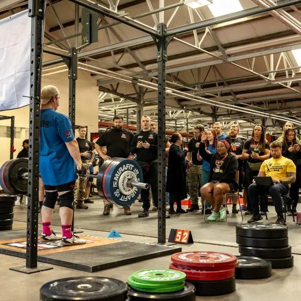 A powerlifter prepares to deadlift in a gym, surrounded by a focused crowd. Weight plates and equipment create an intense, competitive atmosphere.
