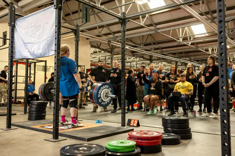 A powerlifter prepares to deadlift in a gym, surrounded by a focused crowd. Weight plates and equipment create an intense, competitive atmosphere.