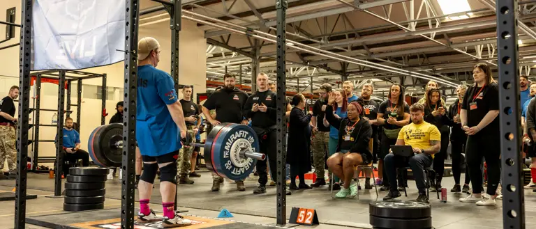 A powerlifter prepares to deadlift in a gym, surrounded by a focused crowd. Weight plates and equipment create an intense, competitive atmosphere.