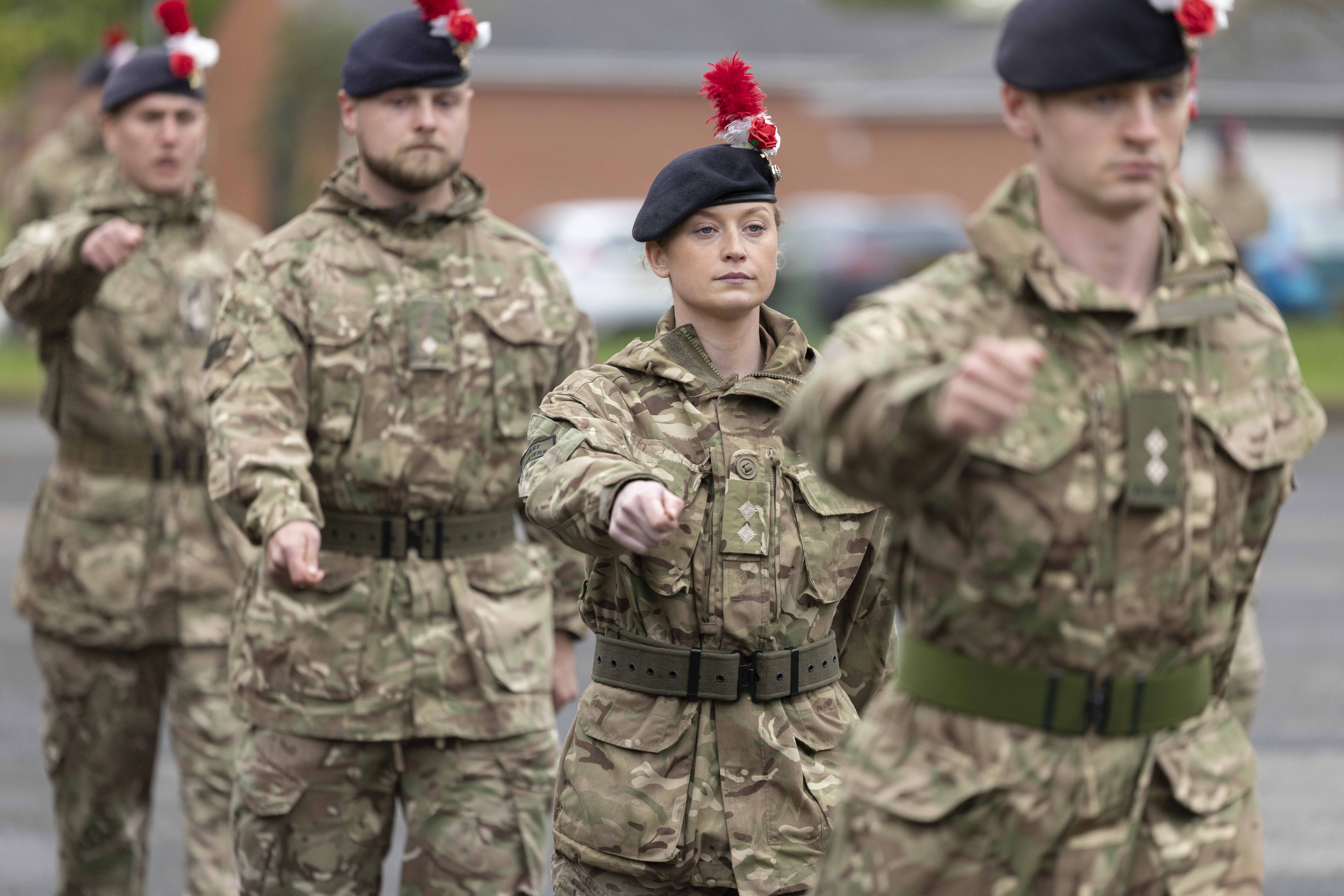 Four soldiers march in camouflage uniforms. On their headdress is a hackle, a white rose and a red rose.