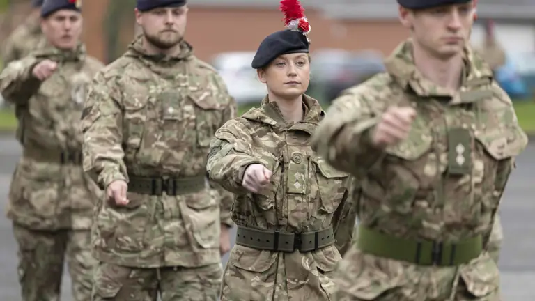 Four soldiers march in camouflage uniforms. On their headdress is a hackle, a white rose and a red rose.