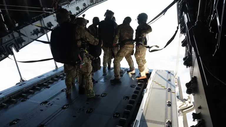 A group of five people in military gear prepare for a parachute jump from an aircraft over the ocean. They are equipped with helmets and harnesses, standing near the open rear door. The vast sea is visible in the background.