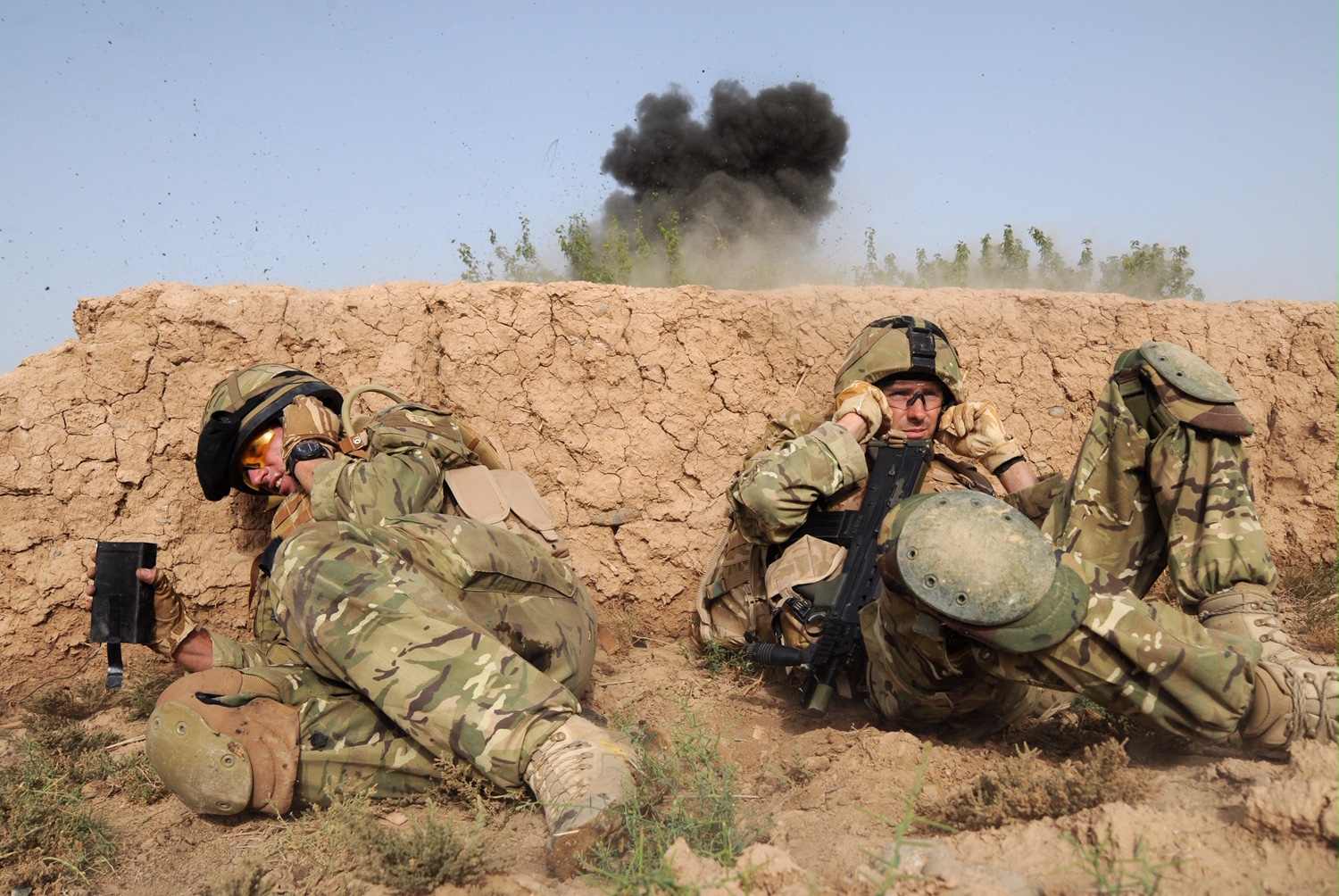 Two soldiers in camouflage uniforms take cover in the foreground from an explosion in the background.