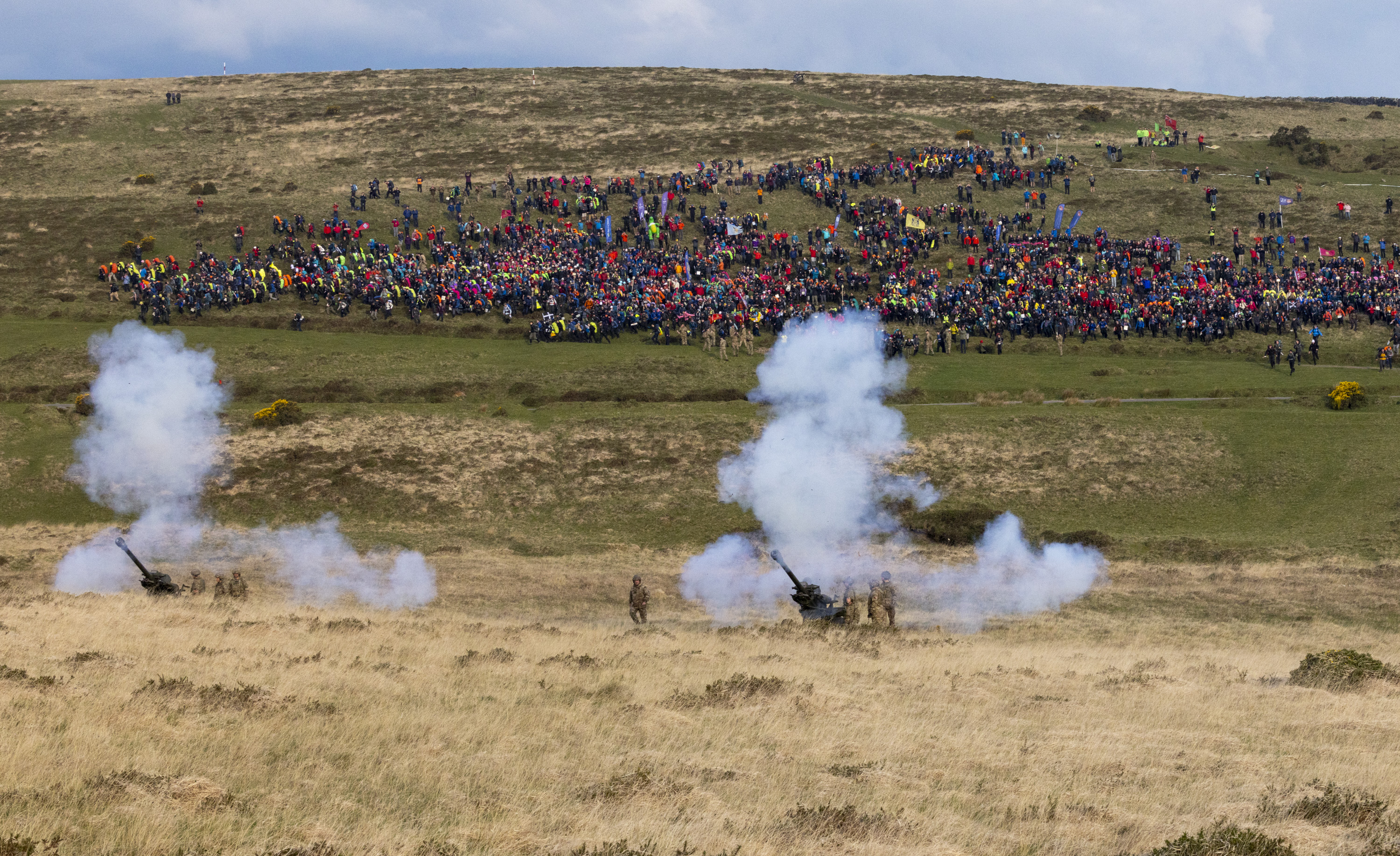 Cannons emit smoke on a grassy field, with a distant crowd of colorful spectators on a hill. The scene conveys energy and excitement.