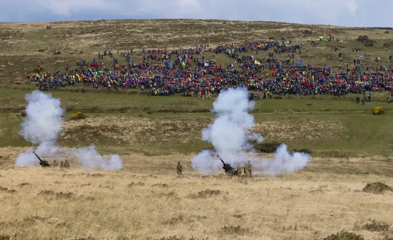 Cannons emit smoke on a grassy field, with a distant crowd of colorful spectators on a hill. The scene conveys energy and excitement.