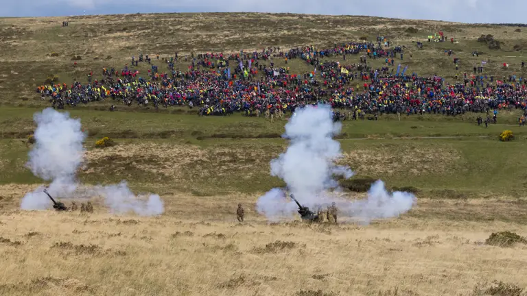 Cannons emit smoke on a grassy field, with a distant crowd of colorful spectators on a hill. The scene conveys energy and excitement.