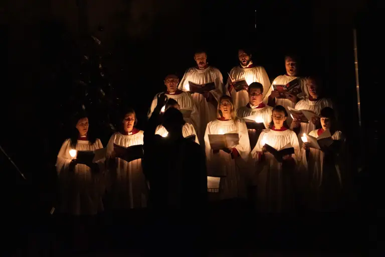 Choir members in white robes singing by candlelight in a dark setting with a conductor in front.
