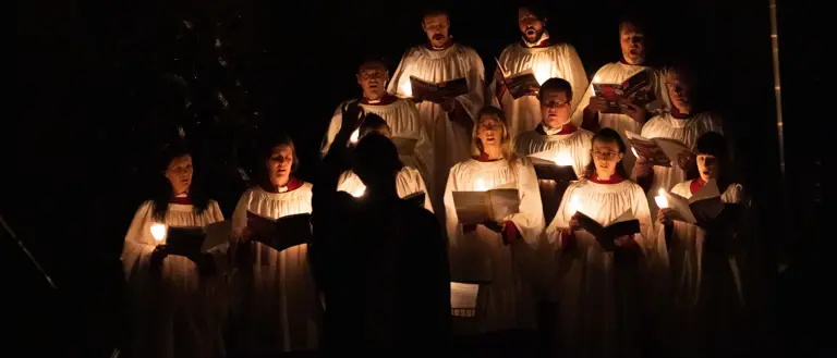 Choir members in white robes singing by candlelight in a dark setting with a conductor in front.