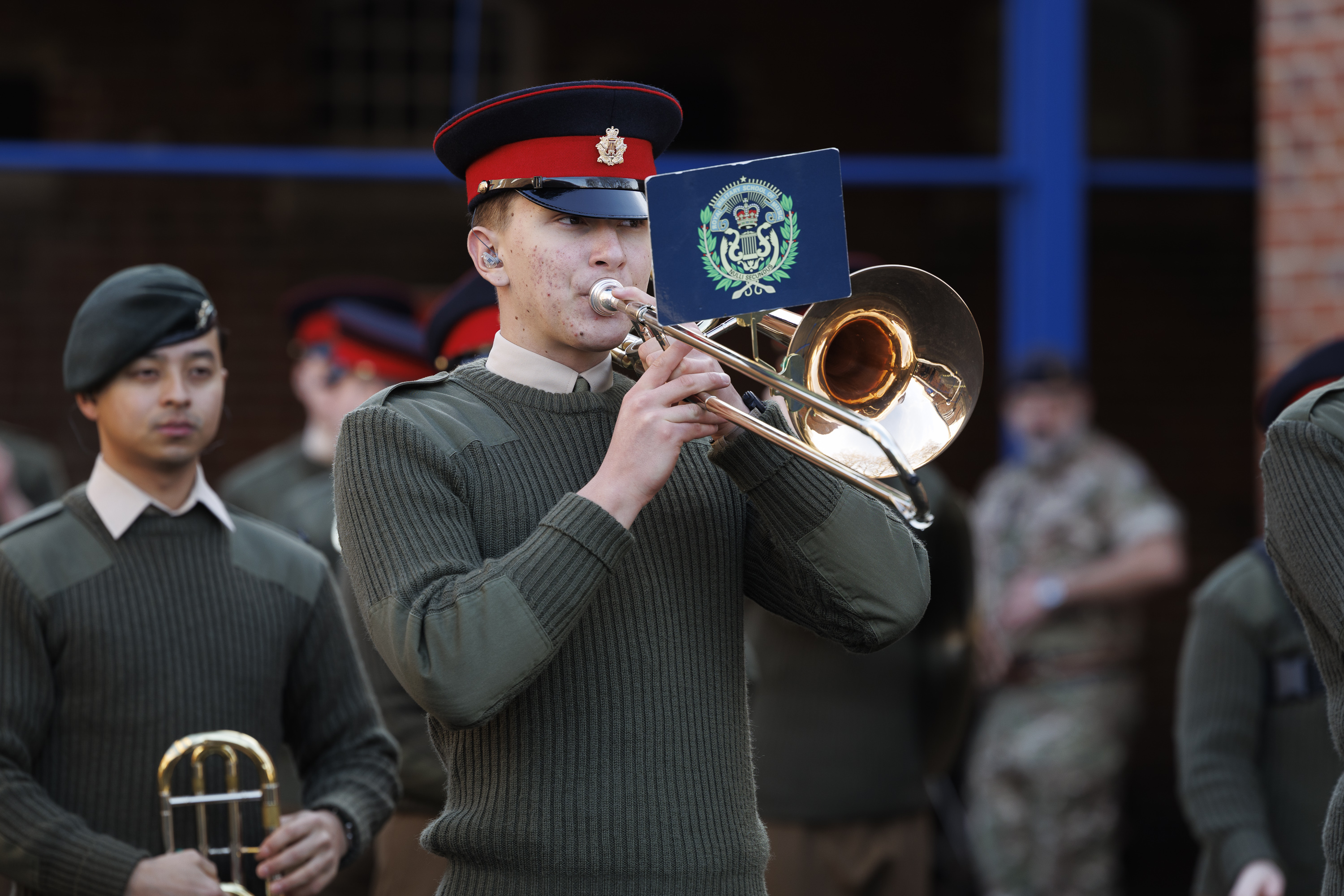 A uniformed band member plays a trombone with a music sheet attached, wearing a red and black cap, surrounded by others in green uniforms, exuding formality.