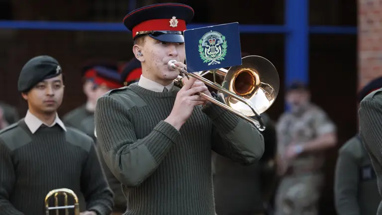 A uniformed band member plays a trombone with a music sheet attached, wearing a red and black cap, surrounded by others in green uniforms, exuding formality.