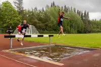 Participants in a track event are seen leaping over a water jump on an outdoor track.