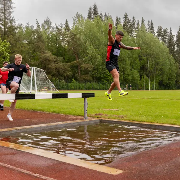 Participants in a track event are seen leaping over a water jump on an outdoor track.