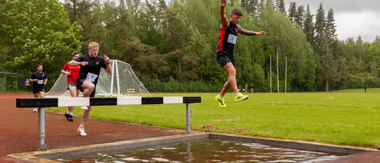 Participants in a track event are seen leaping over a water jump on an outdoor track.