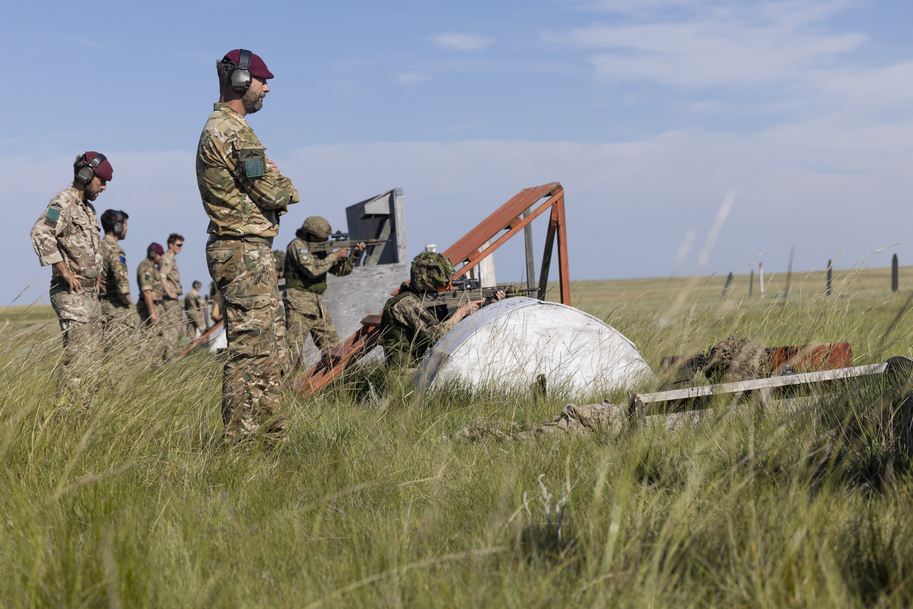 A group of military personnel stands and crouches in a grassy field, engaged in an operation involving a large white object with orange supports. Some are wearing headsets and camouflage uniforms. The sky is clear and blue.
