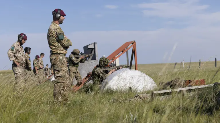 A group of military personnel stands and crouches in a grassy field, engaged in an operation involving a large white object with orange supports. Some are wearing headsets and camouflage uniforms. The sky is clear and blue.