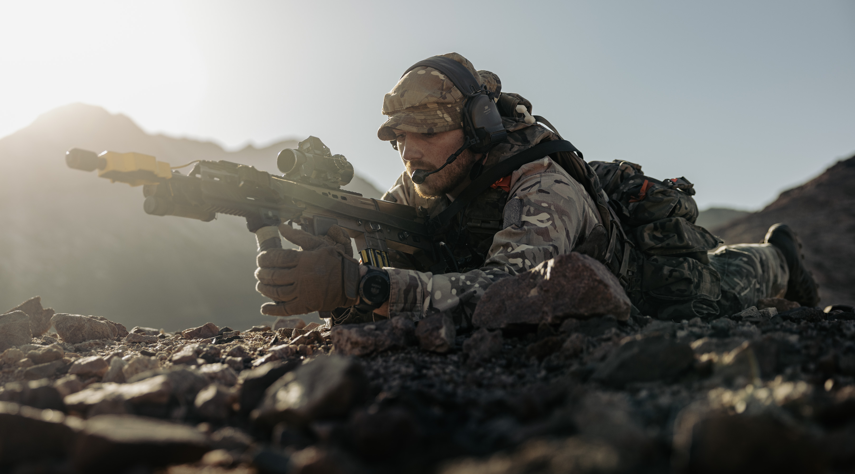 Soldier in camouflage gear lying prone on rocky terrain aiming a scoped rifle during daylight.