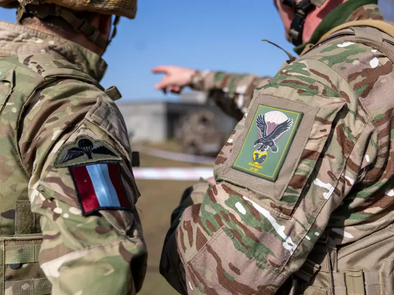 Two soldiers in camouflage uniforms with flashes on their sleeves, one pointing towards a distant structure.