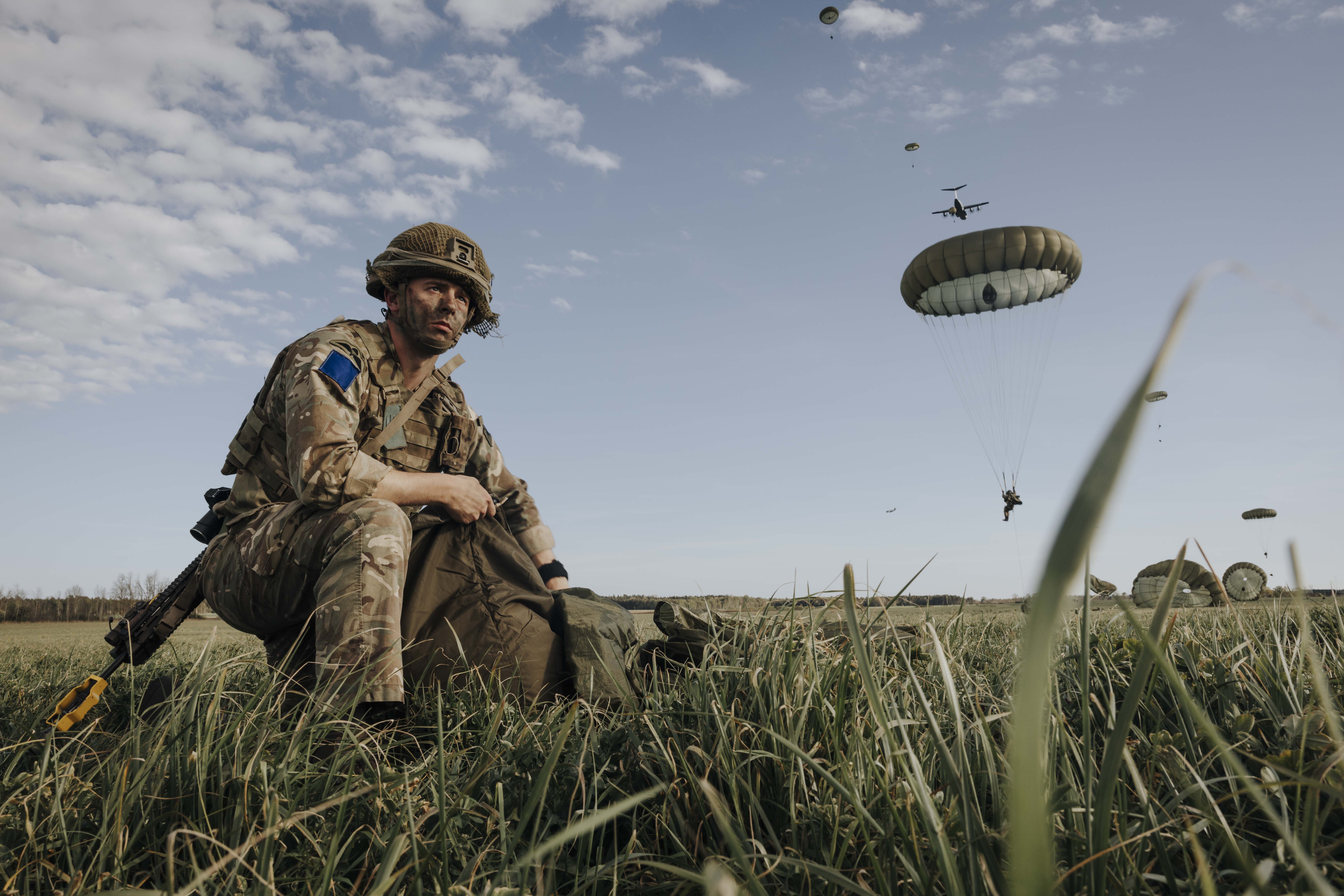 Paratrooper in combat camouflage uniform and helmet packs away his parachute after jumping from a United States A400 aircraft. The air craft is seen in the sky in the distance with other paratroopers in mid jump in the distance. 
