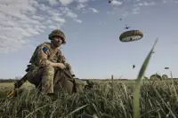 Paratrooper in combat camouflage uniform and helmet packs away his parachute after jumping from a United States A400 aircraft. The air craft is seen in the sky in the distance with other paratroopers in mid jump in the distance.