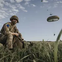 Paratrooper in combat camouflage uniform and helmet packs away his parachute after jumping from a United States A400 aircraft. The air craft is seen in the sky in the distance with other paratroopers in mid jump in the distance.