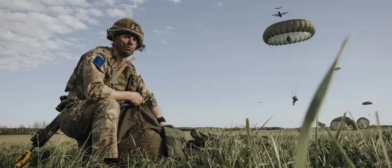 Paratrooper in combat camouflage uniform and helmet packs away his parachute after jumping from a United States A400 aircraft. The air craft is seen in the sky in the distance with other paratroopers in mid jump in the distance.