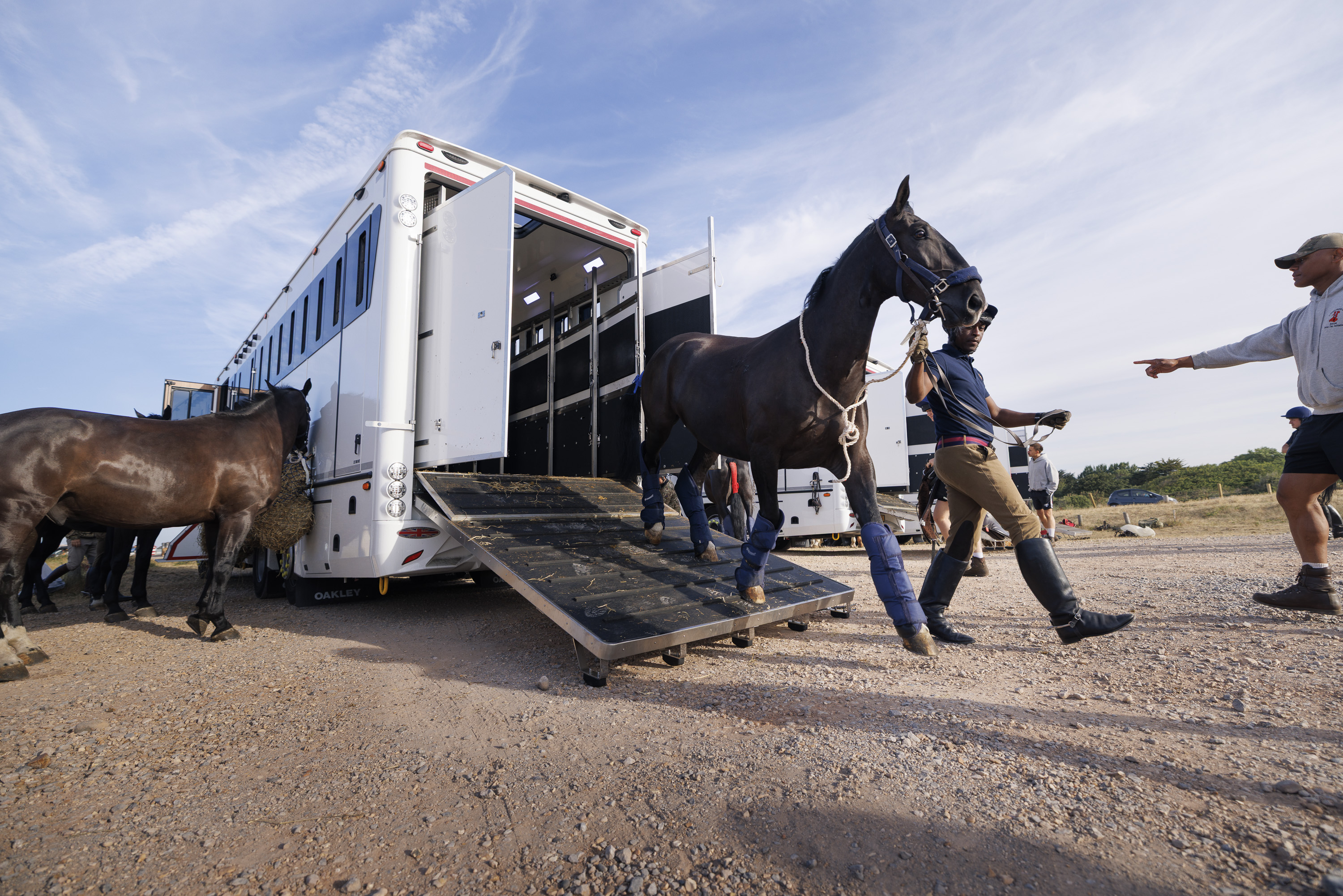 A horse is being led down a ramp from a large, white horse trailer onto a gravel area. Another horse stands nearby. The sky is clear with wispy clouds. Individuals are assisting with the process.