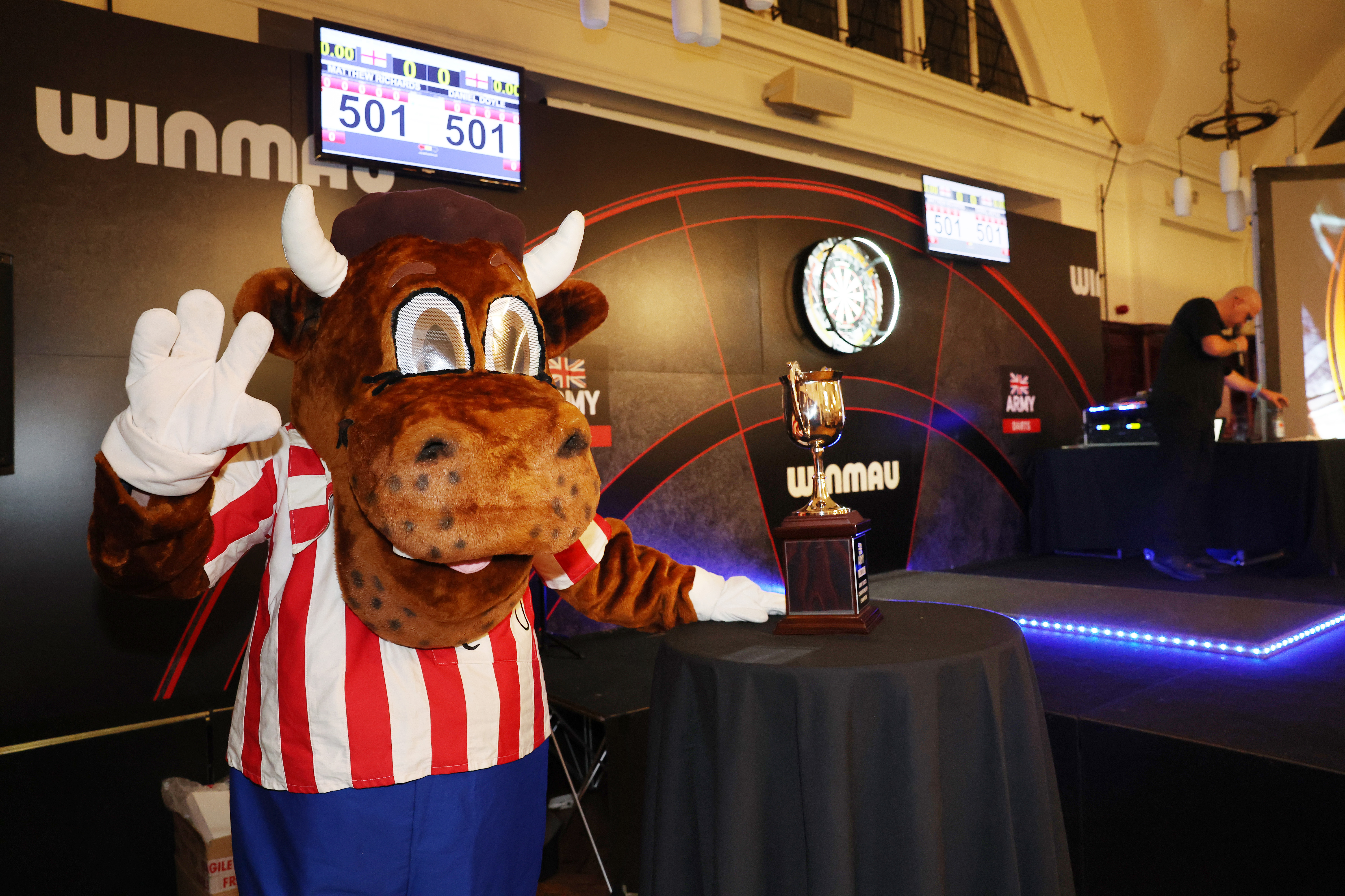 Mascot dressed as a bull in a red and white striped shirt waving next to a trophy on a table at a darts event.