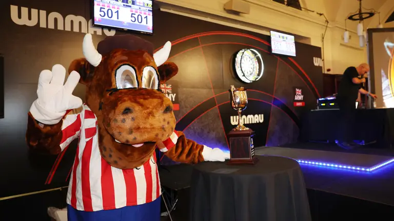 Mascot dressed as a bull in a red and white striped shirt waving next to a trophy on a table at a darts event.