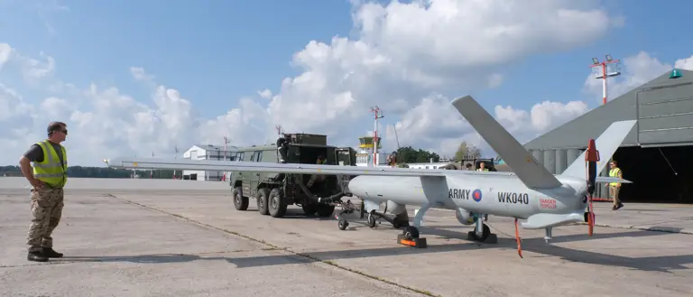 A soldier wearing uniform and a high-vis jacket inspects an unmanned aerial vehicle.